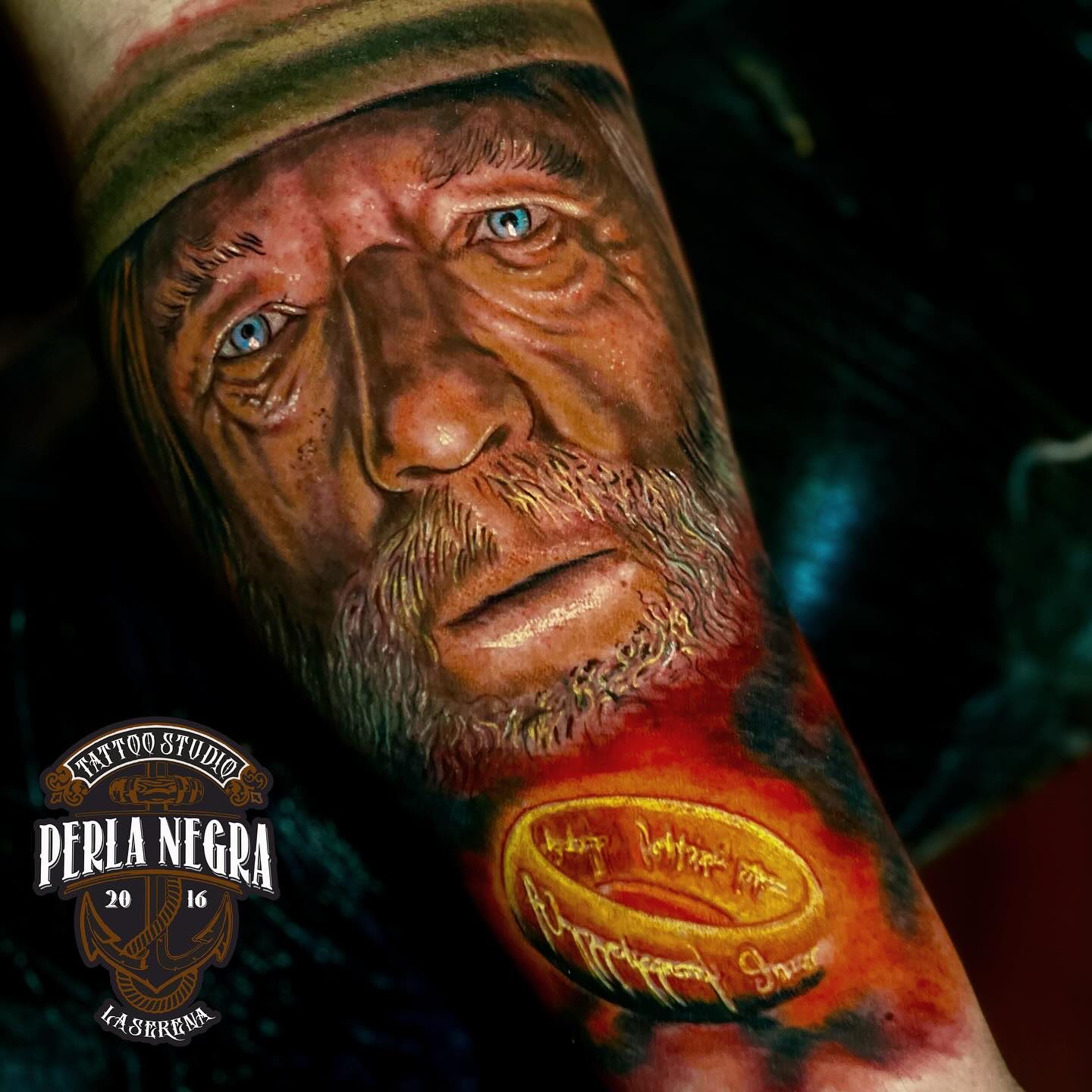 Retrato de un anciano de ojos azules tatuado en un antebrazo, junto a un anillo dorado.